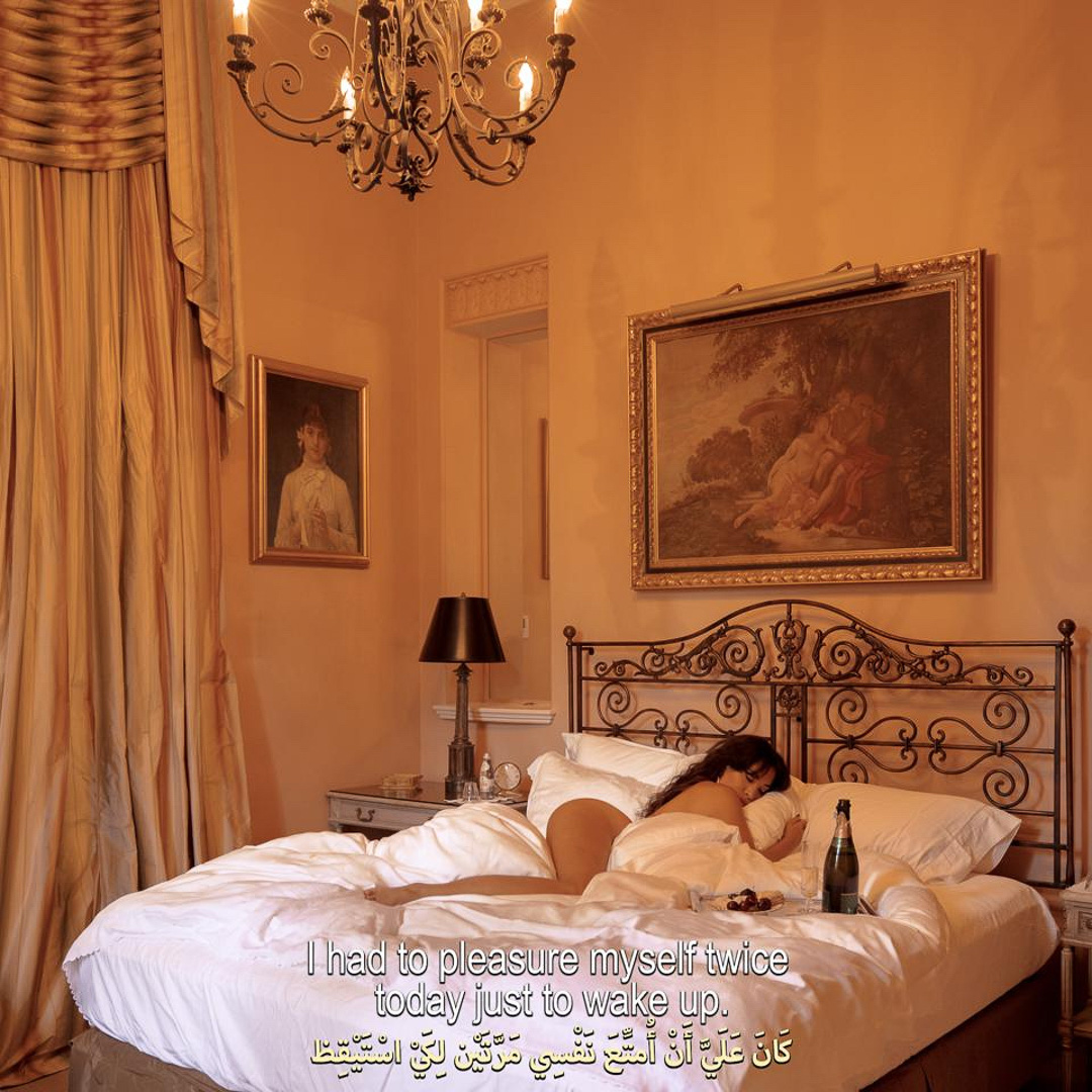A woman lying in an ornate wrought iron bed in a warm-toned bedroom with draped curtains, framed art, and a chandelier.