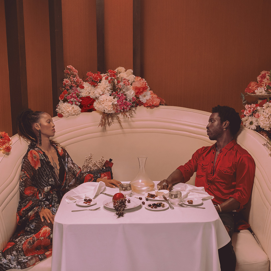 A stylish couple sits at a table with flowers, set against a warm, inviting backdrop. The scene exudes elegance and intimacy.