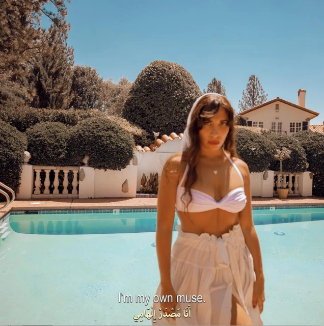 Woman in white bikini top and skirt poses by pool in sunny backyard. House and green bushes create a lush background under a blue sky.