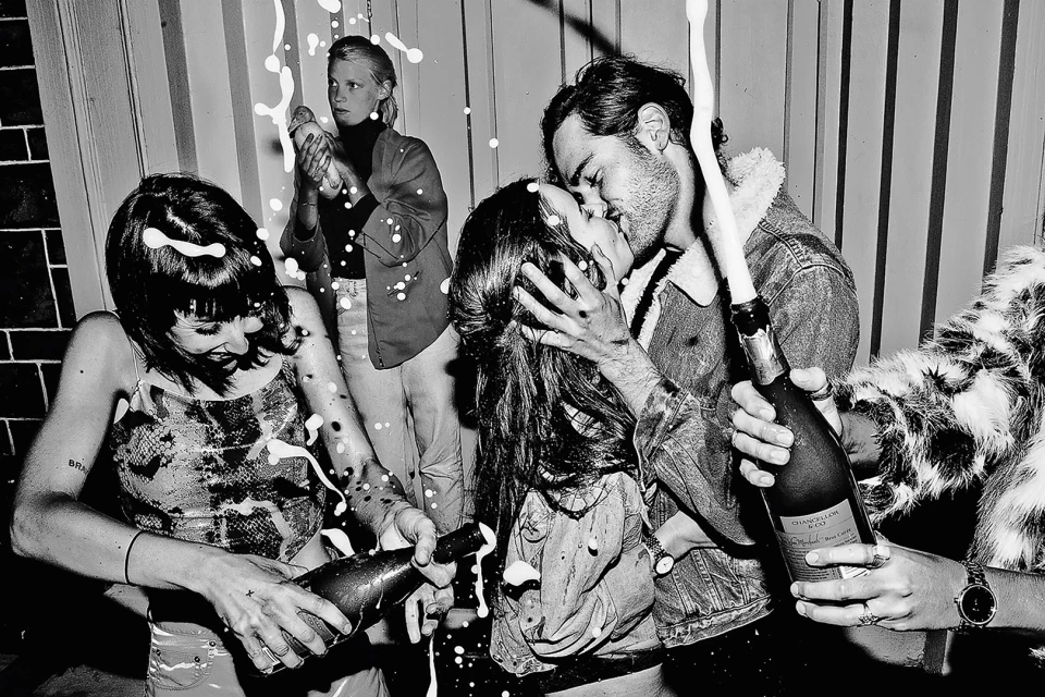 Monochrome photo captures two women opening champagne bottles, while a couple kisses in the background amidst white foam spray.