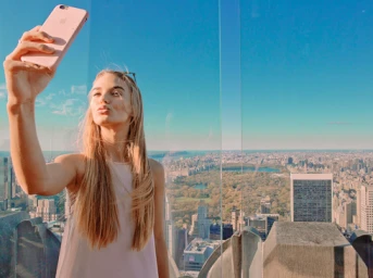 Blonde woman taking a selfie with a pink smartphone against an expansive city skyline backdrop with a large park