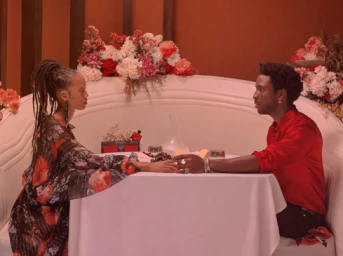 Man in red shirt and woman in floral dress seated across a white table holding hands in a restaurant booth by Sarah Bahbah.