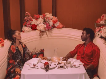 A man and a woman seated at opposite sides of a white-clothed restaurant table with floral arrangements behind them, photographed by Sarah Bahbah.