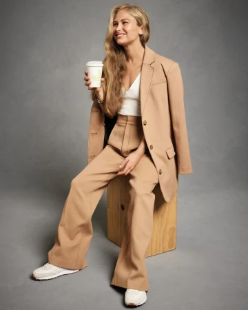 Woman in a tan suit sits, holding a coffee cup. The outfit includes white sneakers, a white top, and blonde hair. Neutral studio setting.