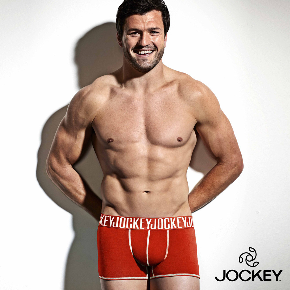 Athletic adult male poses shirtless wearing branded underwear against a white background with subtle shadows and studio lighting.