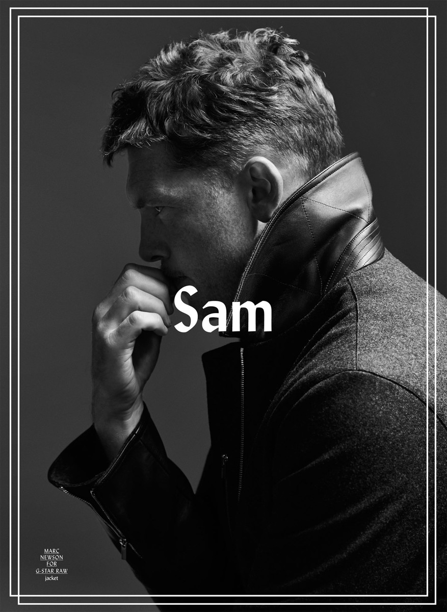 Portrait of a contemplative adult male in a dark jacket, gazing downward with a hand near his face, against a neutral gray backdrop.