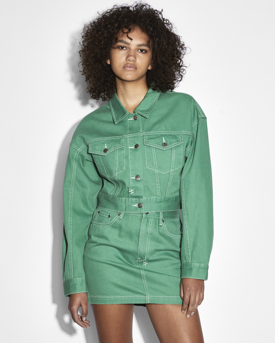 A curly-haired female model poses in a studio wearing a green denim jacket and skirt ensemble with white contrast stitching.