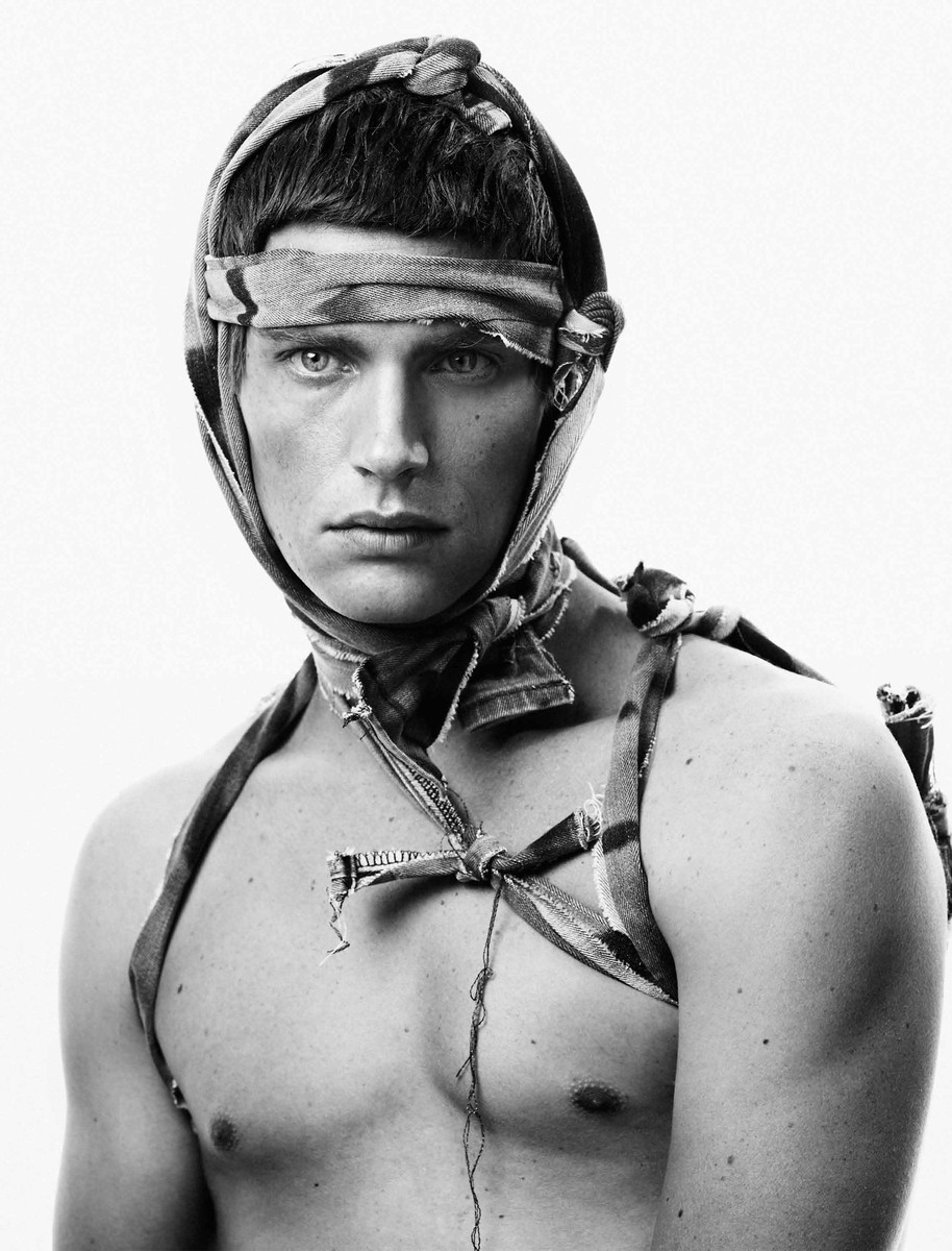Black and white portrait of a man with bare chest, wearing a torn and tied scarf as headwear and harness, against a bright white backdrop.