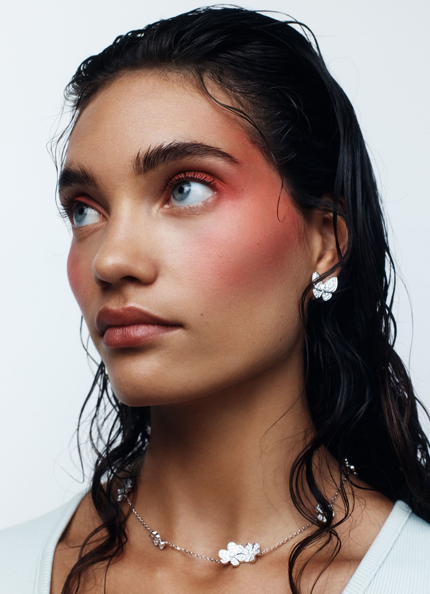 Close-up shot of a woman with blush makeup wearing jewelry with flower details against a white background in a beauty shot.
