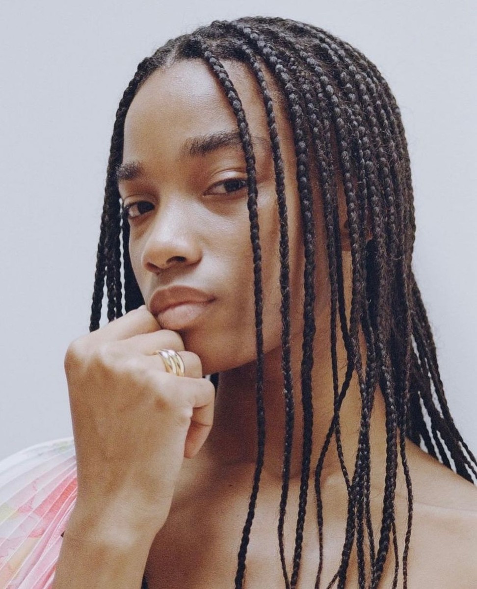 Close-up studio portrait of a young adult woman with long dark braids, resting her chin on her hand and wearing rings.
