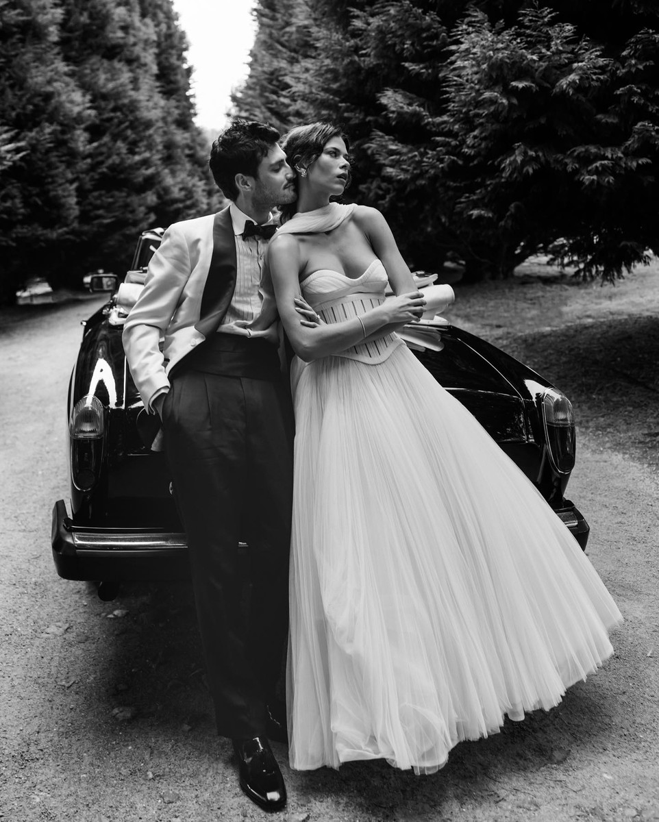 Black and white outdoor shot of a stylish couple in formalwear posing next to a vintage car with trees lining the background.