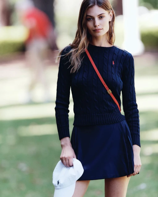 A young adult woman poses outdoors wearing a navy cable knit sweater, matching skirt, and holding a white baseball cap.