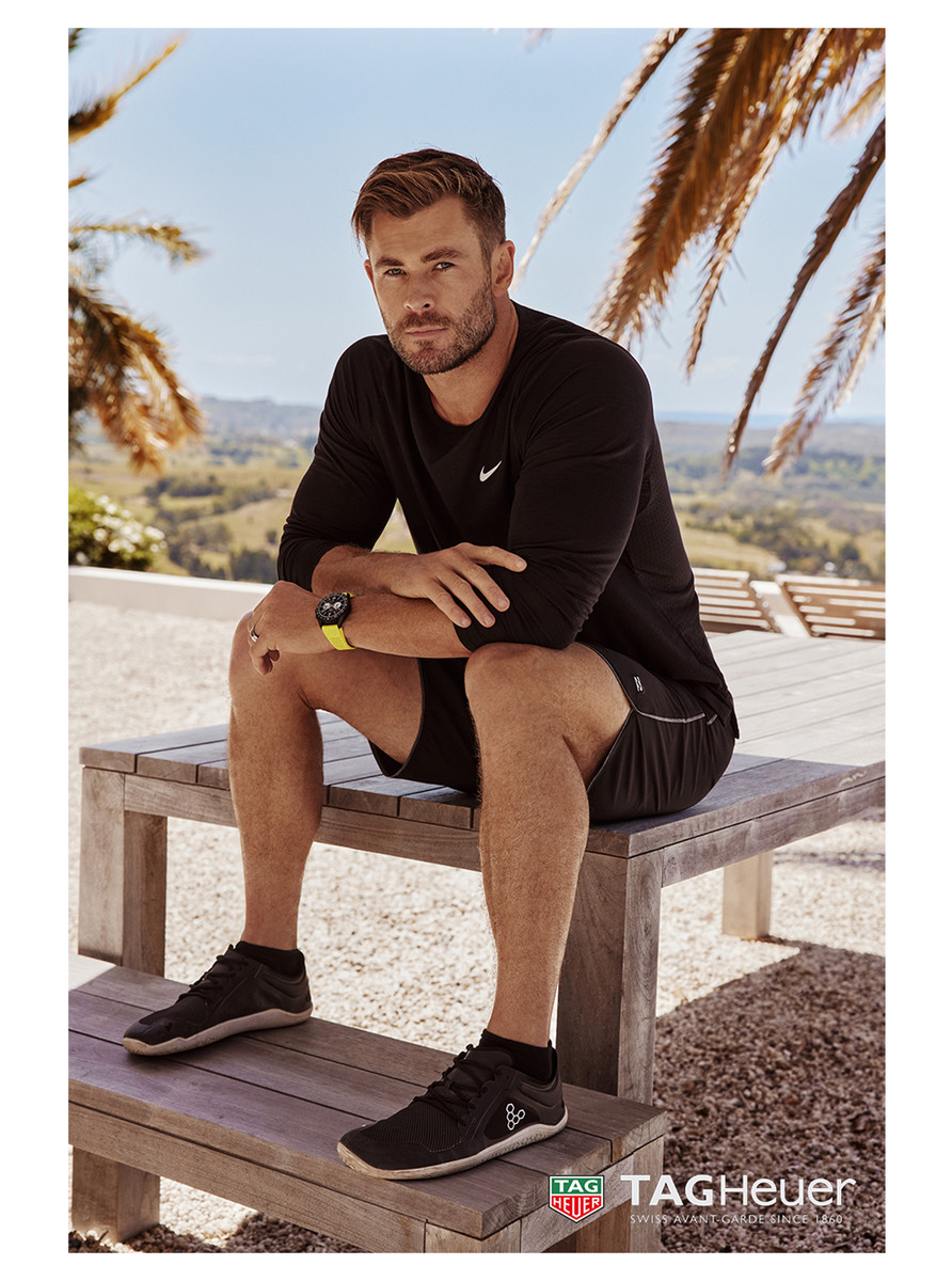 Adult male model, Chris Hemsworth, poses on a wooden bench in activewear, with palm trees and an outdoor environment in the background.