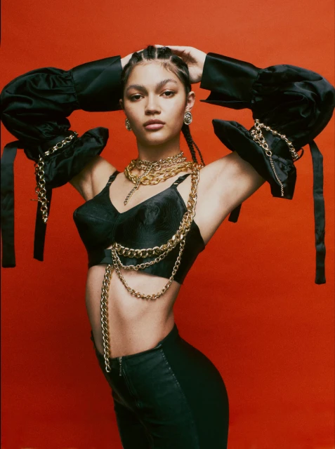 Portrait of an adult woman in a black bra top and pants with gold chain accessories against a vibrant red backdrop in a photo studio.