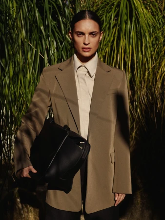 Woman in a tailored taupe blazer and collared shirt holding a black handbag amidst tropical foliage
