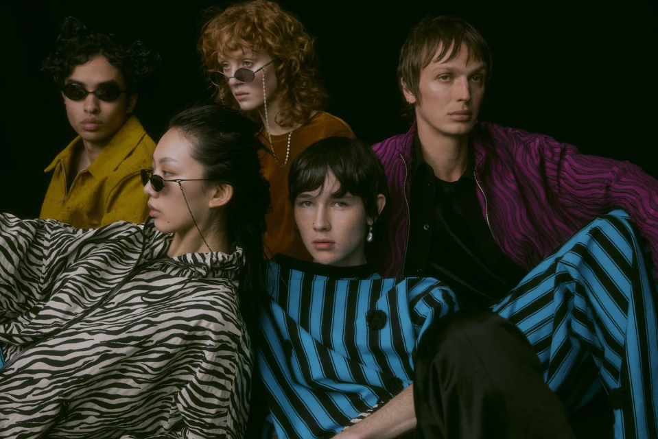 Group of five fashionably dressed young adults in animal prints and stripes posing against a dark background, wearing sunglasses.