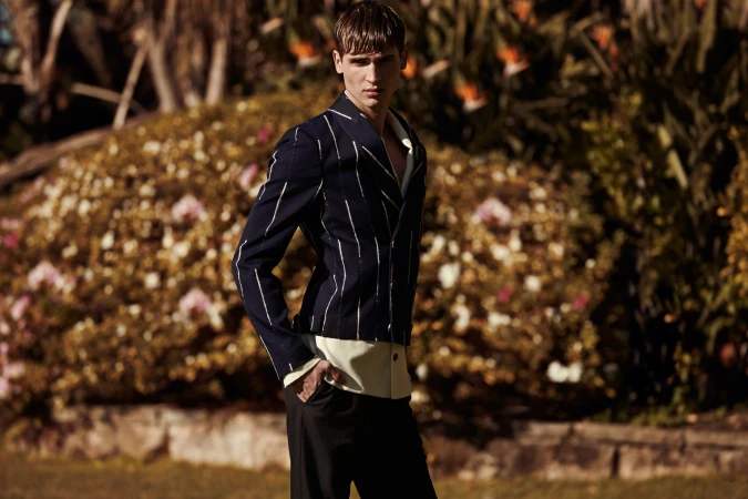 Male model in pinstripe blazer and dark trousers posing outdoors with garden foliage background