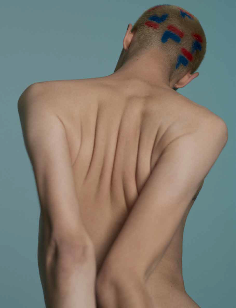 Nude person's back and head with painted symbols on a light blue background in studio lighting showcasing skin tones and artistic design.