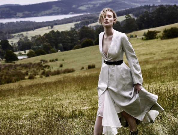 Model with short blonde hair wearing a long white coat walking across a lush green hillside with a lake in the background.