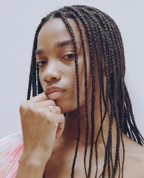 Portrait of a Black woman with long box braids resting her chin on her hand against a plain background