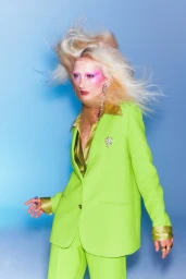 Model with wind-swept blonde hair and pink eyeshadow wearing a lime green suit against a blue backdrop