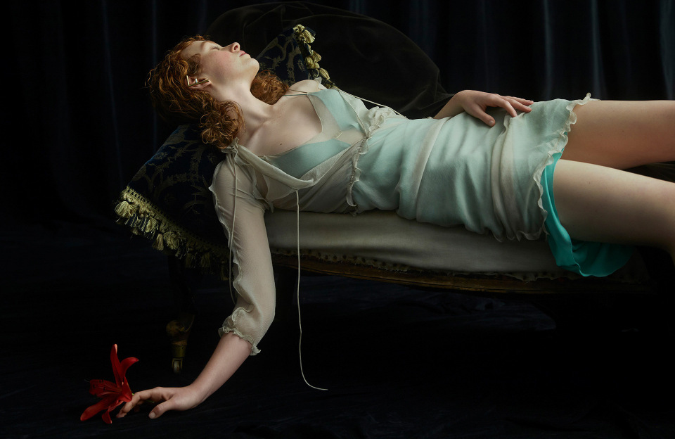 Red-haired woman in a light blue dress lies on a blue patterned pillow with a red flower on a dark blue backdrop in a studio setting.
