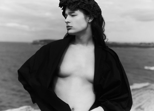 Monochrome portrait of a woman wearing a dark textured headpiece and draped black garment against an ocean backdrop.