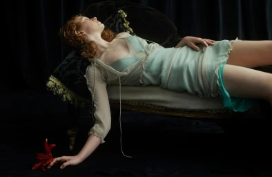 Red-haired woman in a light blue dress lying on an ornate chaise lounge, reaching toward a red lily on a dark floor.