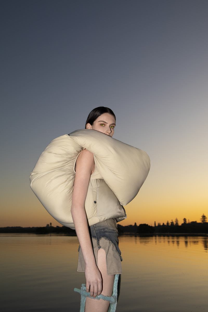 A woman stands at the water's edge at dusk, wearing a beige puffy vest and denim shorts against a gradient sky with the sun setting.
