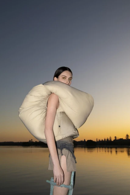 A woman stands at the water's edge at dusk, wearing a beige puffy vest and denim shorts against a gradient sky with the sun setting.