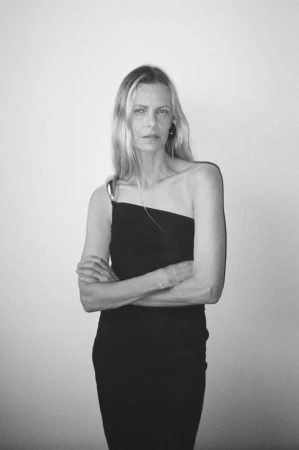 Black and white portrait of a woman with long blonde hair wearing a dark one-shoulder dress with arms crossed against a plain wall