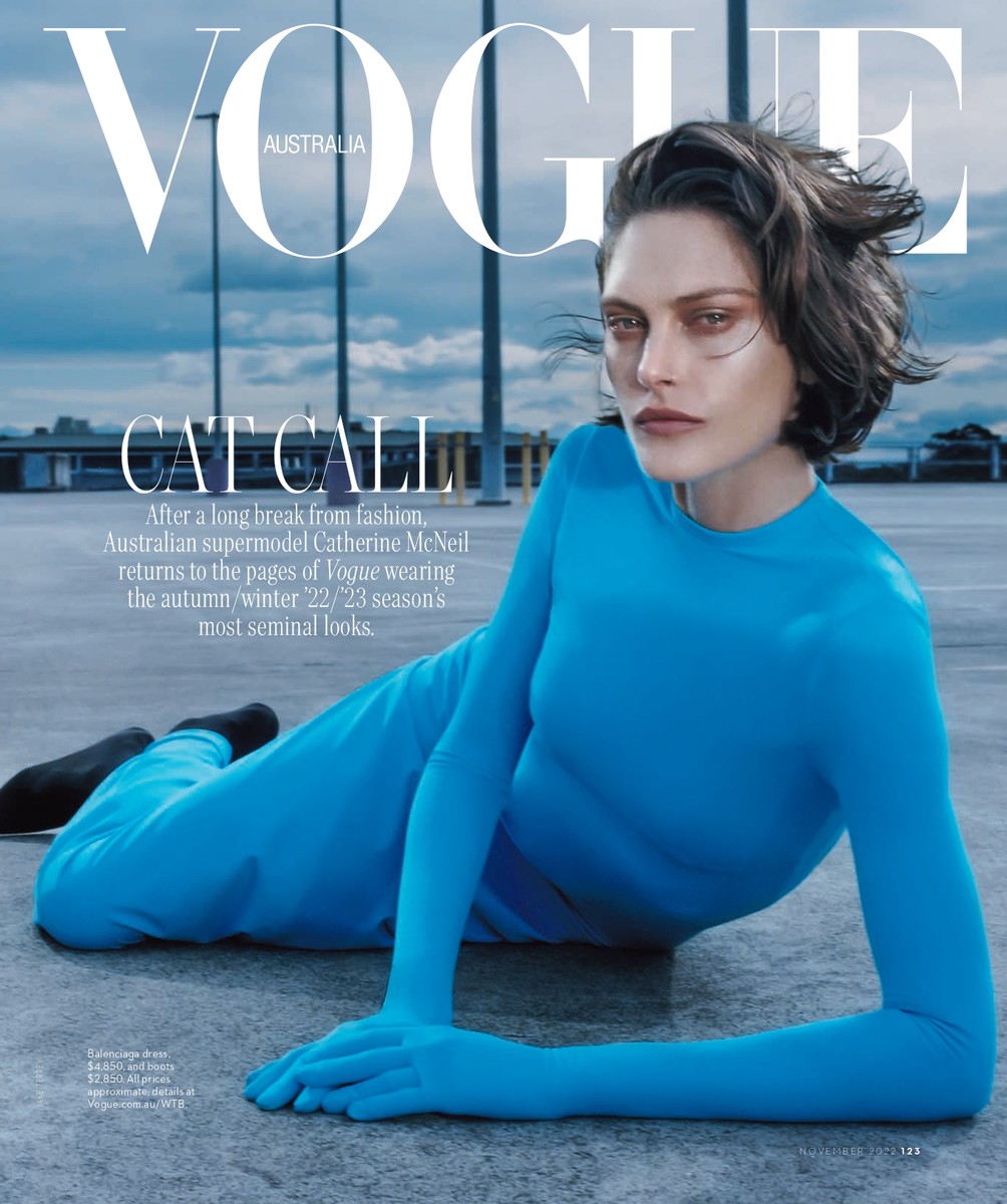 Model Catherine McNeil lying on a rooftop wearing a bright blue longsleeve jumpsuit under an overcast sky.
