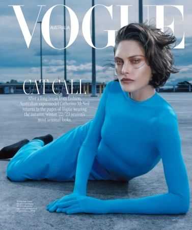 Model Catherine McNeil lying on a rooftop wearing a bright blue longsleeve jumpsuit under an overcast sky.
