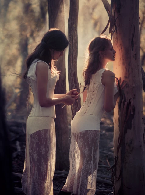 Two women in white lace dresses standing near a tree in a forest, with one tying the other's corset with natural light.