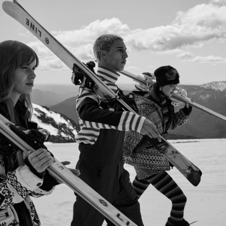 Three people in winter clothing carry skis across snow, featuring mountains and clouds in the background, shot in black and white.