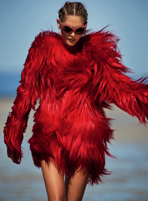 A model in a striking red faux fur coat and decorative sunglasses stands on a beach with the sea and sky in the background.