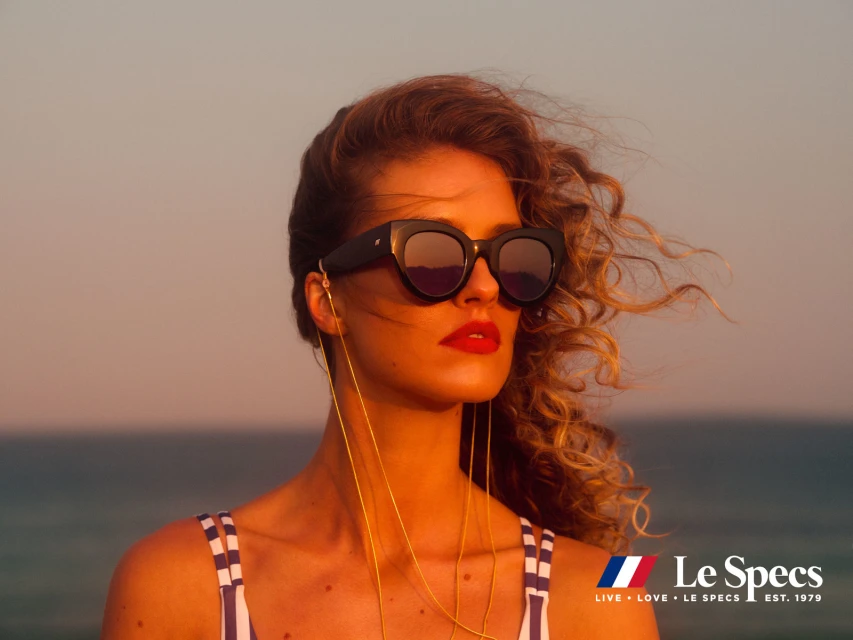 Woman in sunglasses poses against ocean and sky at sunset. She wears a striped top and gold chain; the 'Le Specs' logo is visible.