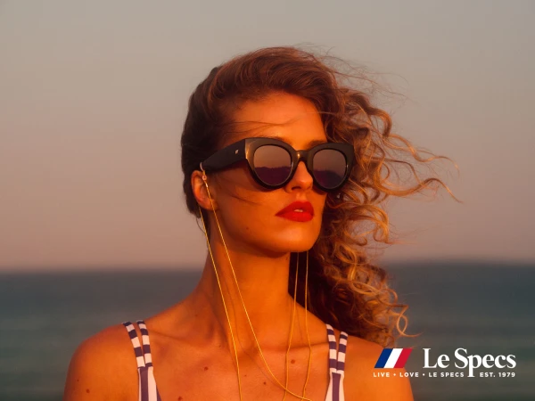 Woman in sunglasses poses against ocean and sky at sunset. She wears a striped top and gold chain; the 'Le Specs' logo is visible.