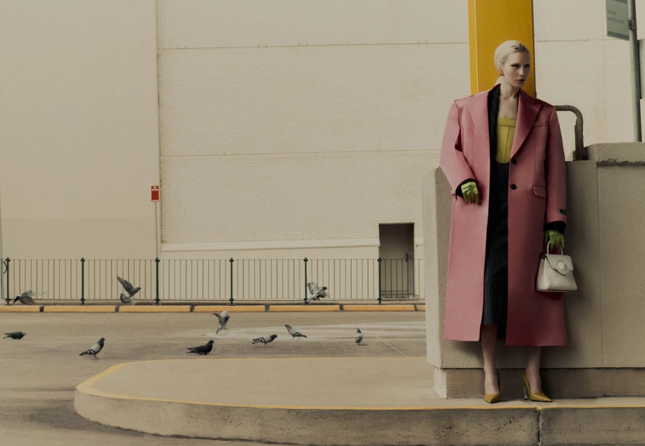 Stylish woman in a pink coat posing outdoors near pigeons, accessorized with a light-colored purse and green gloves. Fashion editorial.