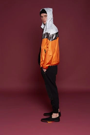 A young adult male is standing in a studio wearing a black beanie, black athletic pants, and a hooded jacket with a color block design.
