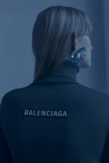 Woman in a navy turtleneck sweater with a prominent 'BALENCIAGA' logo on the back, highlighted by striking blue hues and bold earrings.