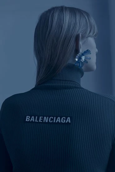 Woman in a navy turtleneck sweater with a prominent 'BALENCIAGA' logo on the back, highlighted by striking blue hues and bold earrings.