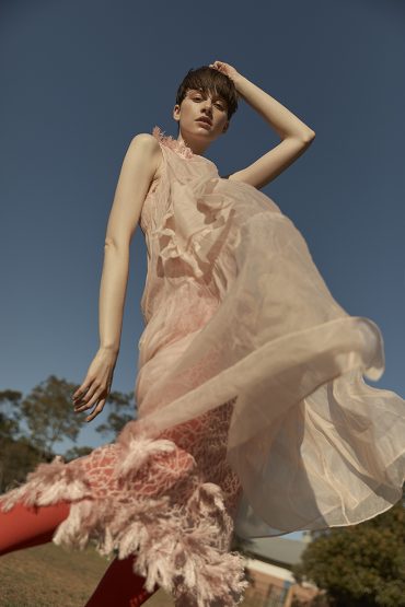 Woman in sheer pink dress poses outdoors against a blue sky. Low angle captures the flowing fabric and her expressive gaze.