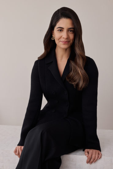 Portrait of a brunette woman in a black jacket and pants sitting against a neutral background in a medium shot with natural lighting.