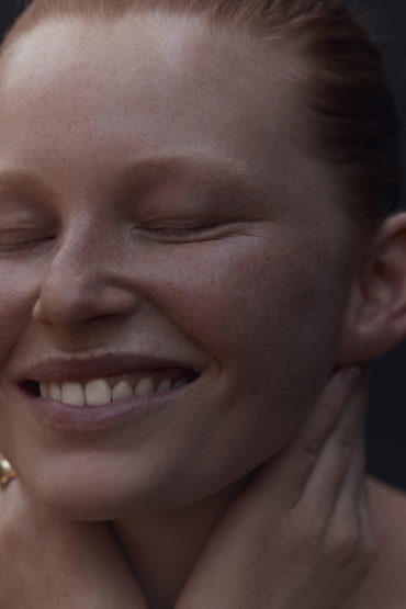 Close-up of a smiling woman with red hair and closed eyes, hands at her neck against a dark background, creating a peaceful and serene portrait.