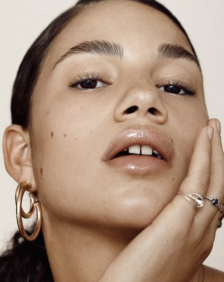 Close-up portrait of a young adult female with a neutral facial expression, glossy lips, and gold hoop earrings against a light background.