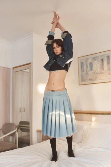 A stylish woman in a denim jacket and skirt is posing on a bed in an indoor bedroom setting with a playful and energetic vibe.
