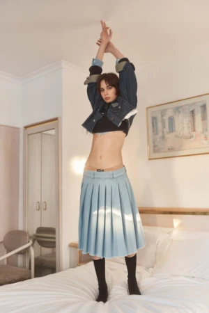 A stylish woman in a denim jacket and skirt is posing on a bed in an indoor bedroom setting with a playful and energetic vibe.