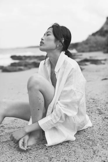 Black and white portrait of an Asian woman in a white shirt sitting on a sandy beach with a blurred rocky coastline in the background.
