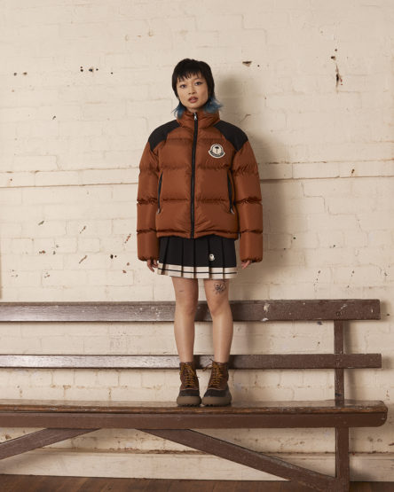 Young woman in a stylish brown puffer jacket with a black and white pleated skirt and brown boots standing in a casual pose.